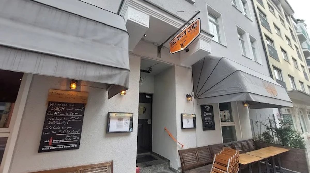 Eingang der Drunken Cow in der Gabelsbergerstraße mit Markise, Tafel und Holztischen vor dem Haus