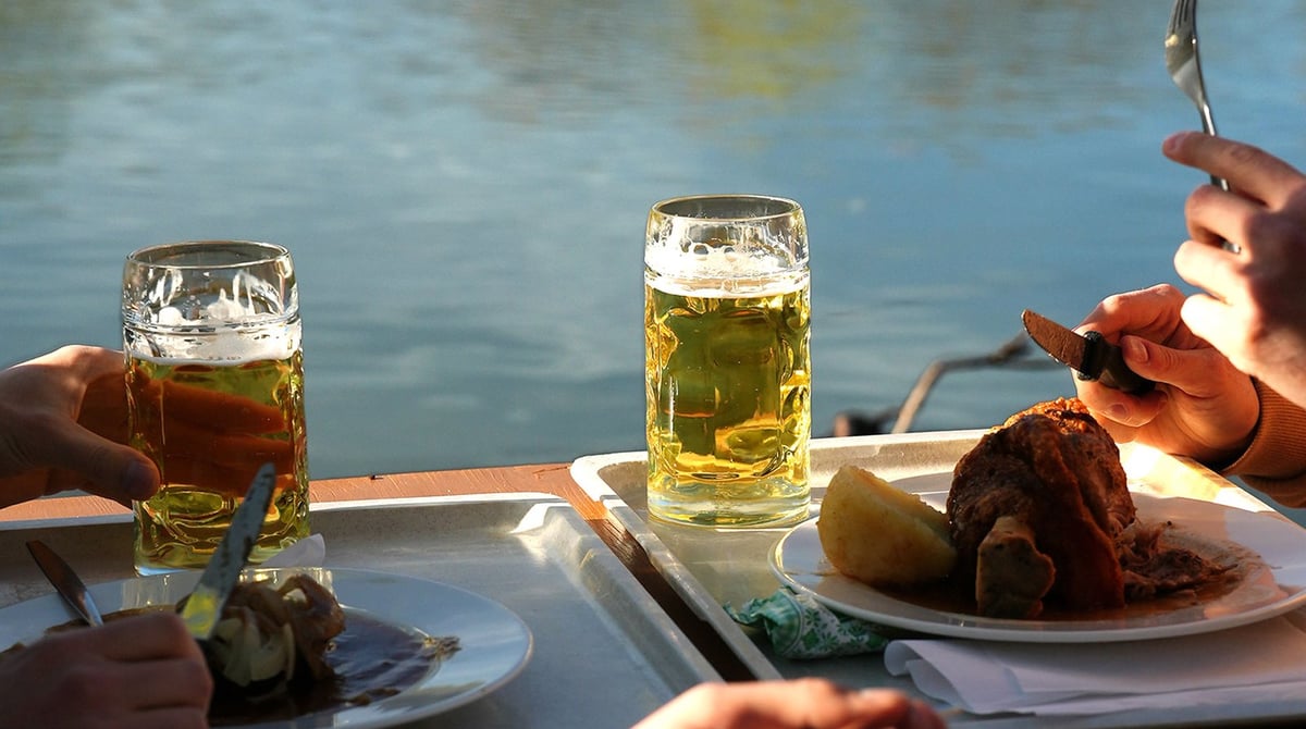 Zwei Gläser Bier und ein Teller mit Braten auf einem Tisch am Wasser