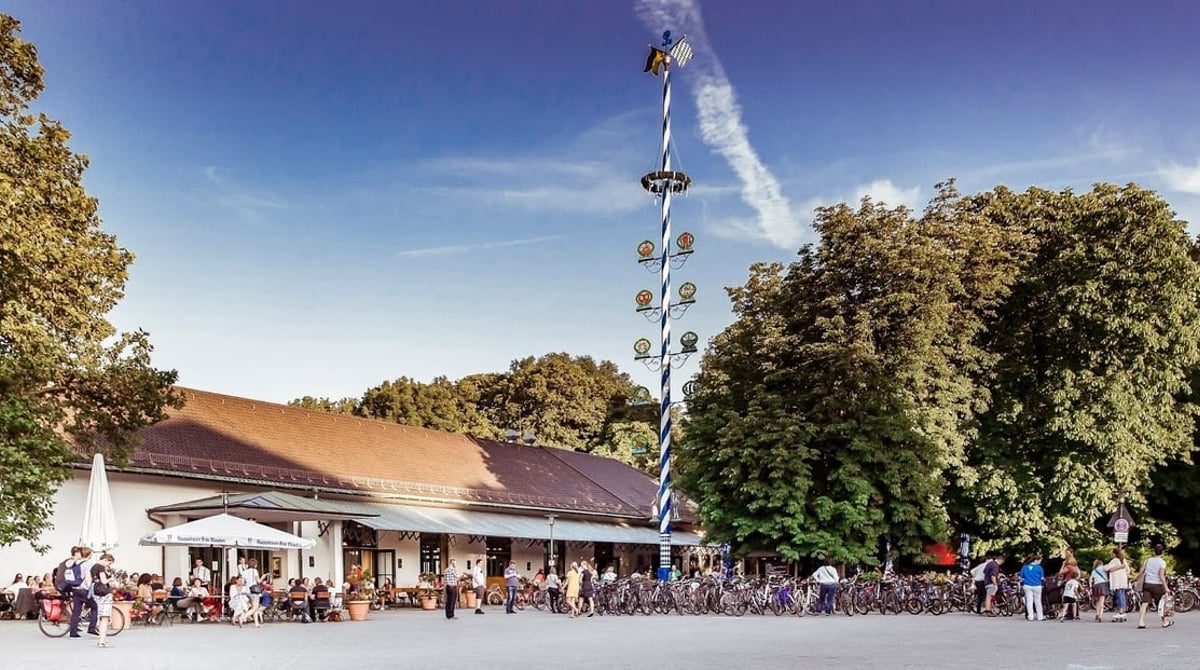 Wirtshaus am Bavariapark mit Maibaum und Biergarten unter hohen Bäumen vor blauem Himmel
