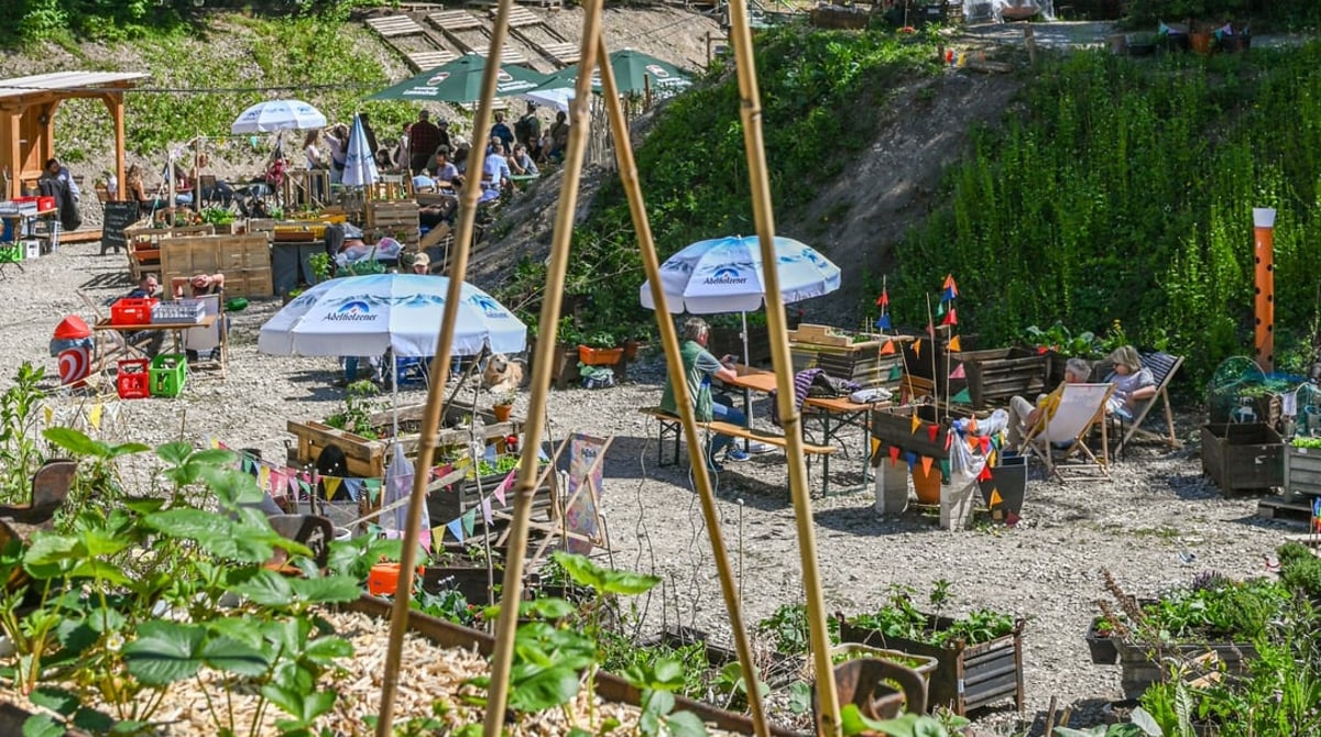 Biergartentische mit Sonnenschirmen, Liegestühlen und Hochbeeten auf dem Kiesgrund der Baugrube, dahinter hohe Bäume