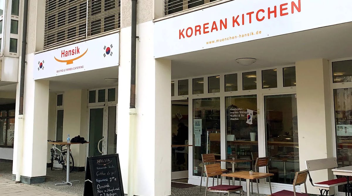 Straßenansicht des Hansik mit den Schildern „Korean Kitchen" und „Bistro & Firmen Catering" und Tischen vor dem Lokal