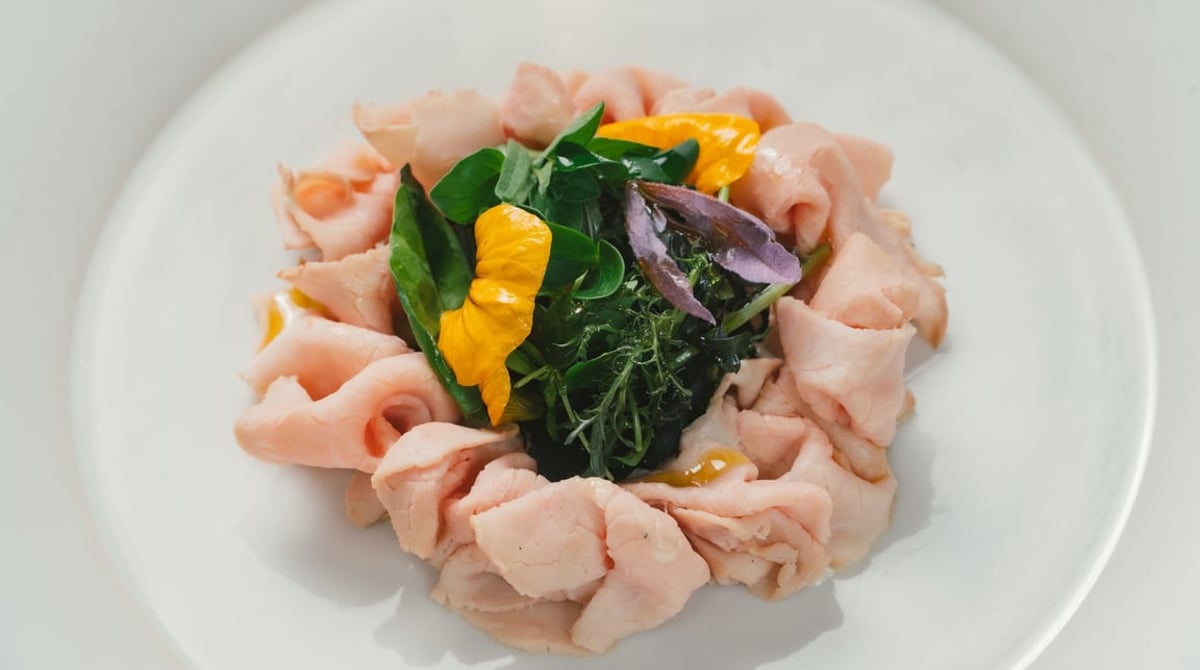Aufgerollte rosa Fleischscheiben mit Kräutersalat und gelben Blüten auf einem weißen Teller
