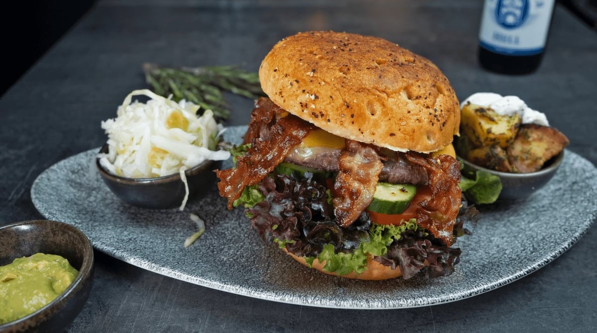 Burger mit Bacon und Salat, dazu Krautsalat, Guacamole und Kartoffeln auf einem Teller