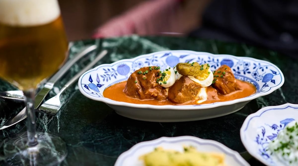 Fleischgericht in oranger Sauce mit Ei auf blau-weißem Geschirr, links ein Glas Bier