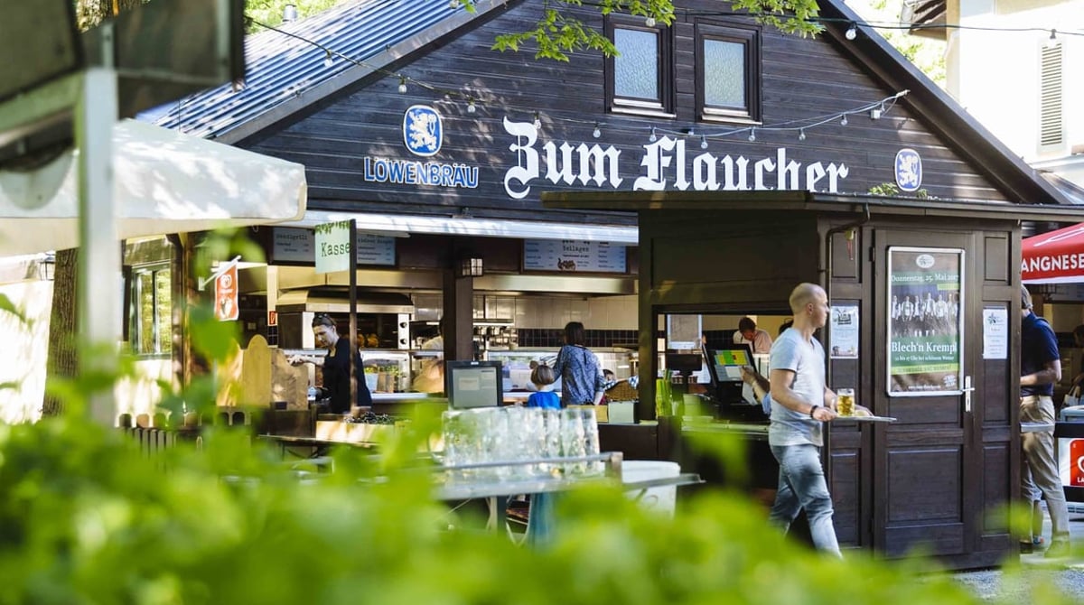 Schänke des Biergartens Zum Flaucher mit Löwenbräu-Schild, davor grünes Laub
