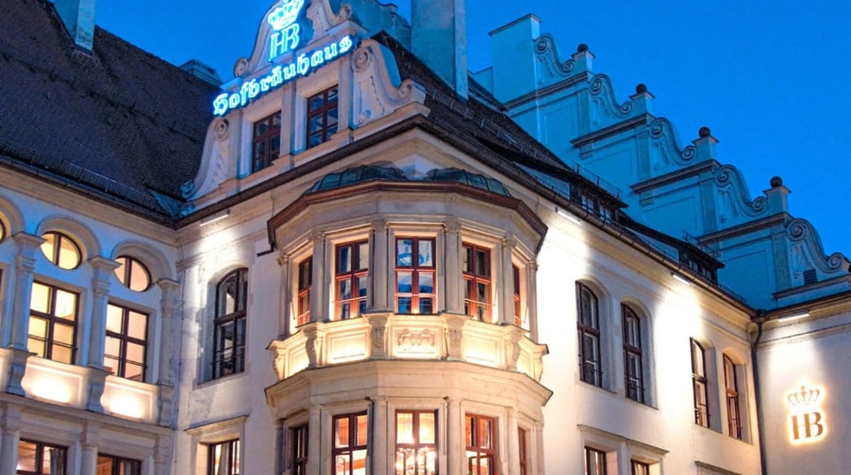 Das abends beleuchtete Hofbräuhaus am Platzl mit Erker, Leuchtschrift und Blumenschmuck vor blauem Himmel