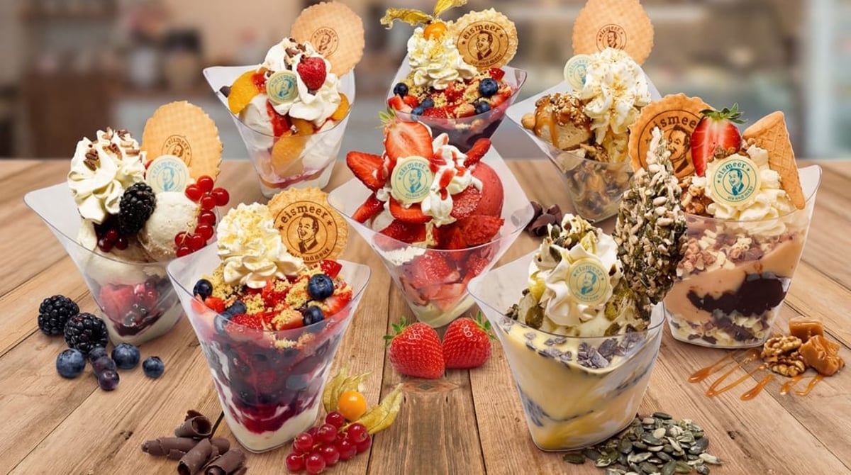 Sieben Eisbecher mit Sahne, Früchten, Waffeltalern und Nüssen auf einem Holztisch