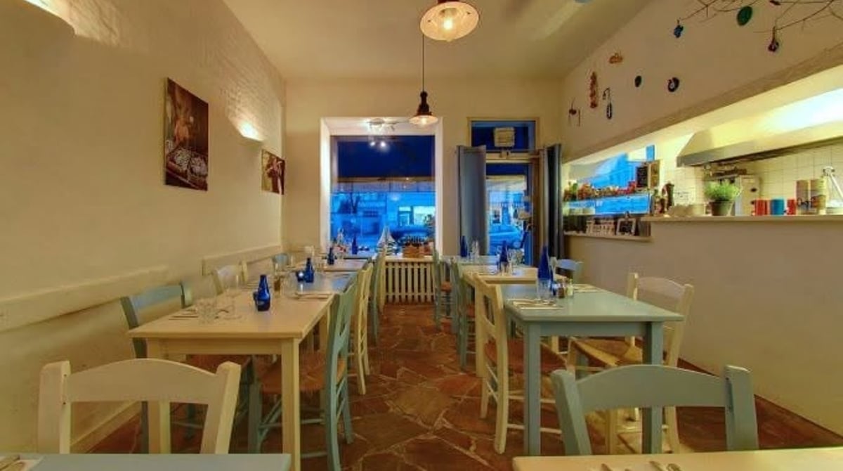 Gastraum des Kreta Grill mit blau-weiß eingedeckten Holztischen, Steinboden und Blick zur Fensterfront