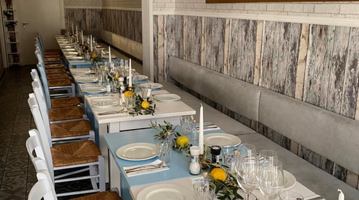 Gastraum des Kreta Grill mit langer gedeckter Tafel, weißer Ziegelwand und Strohstühlen