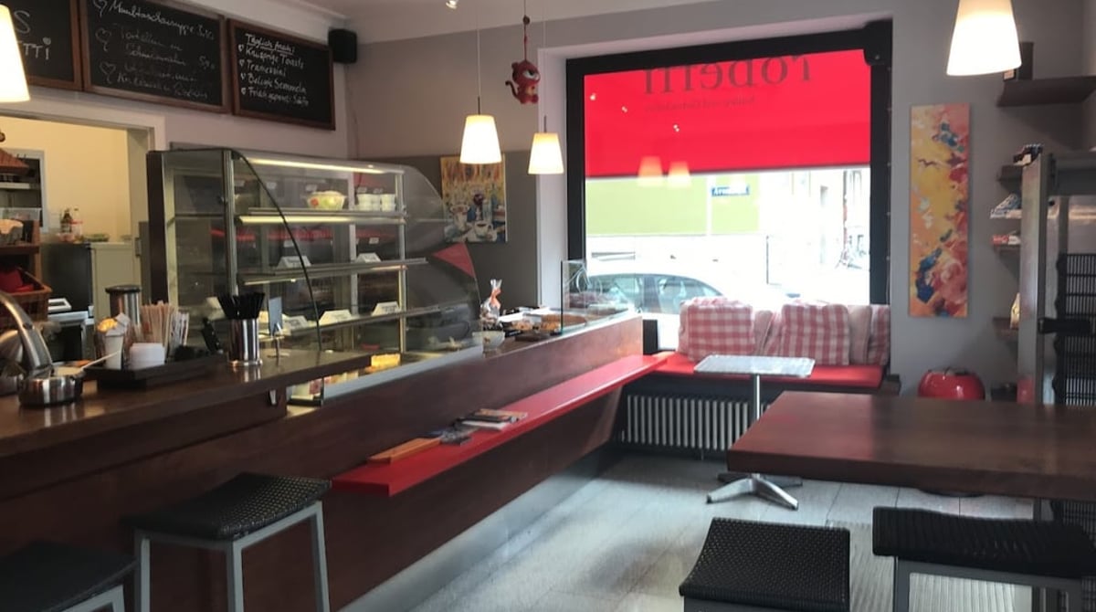 Gastraum des Café Robetti mit Holztheke, Vitrine, Barhockern und roter Fensterbank mit karierten Kissen