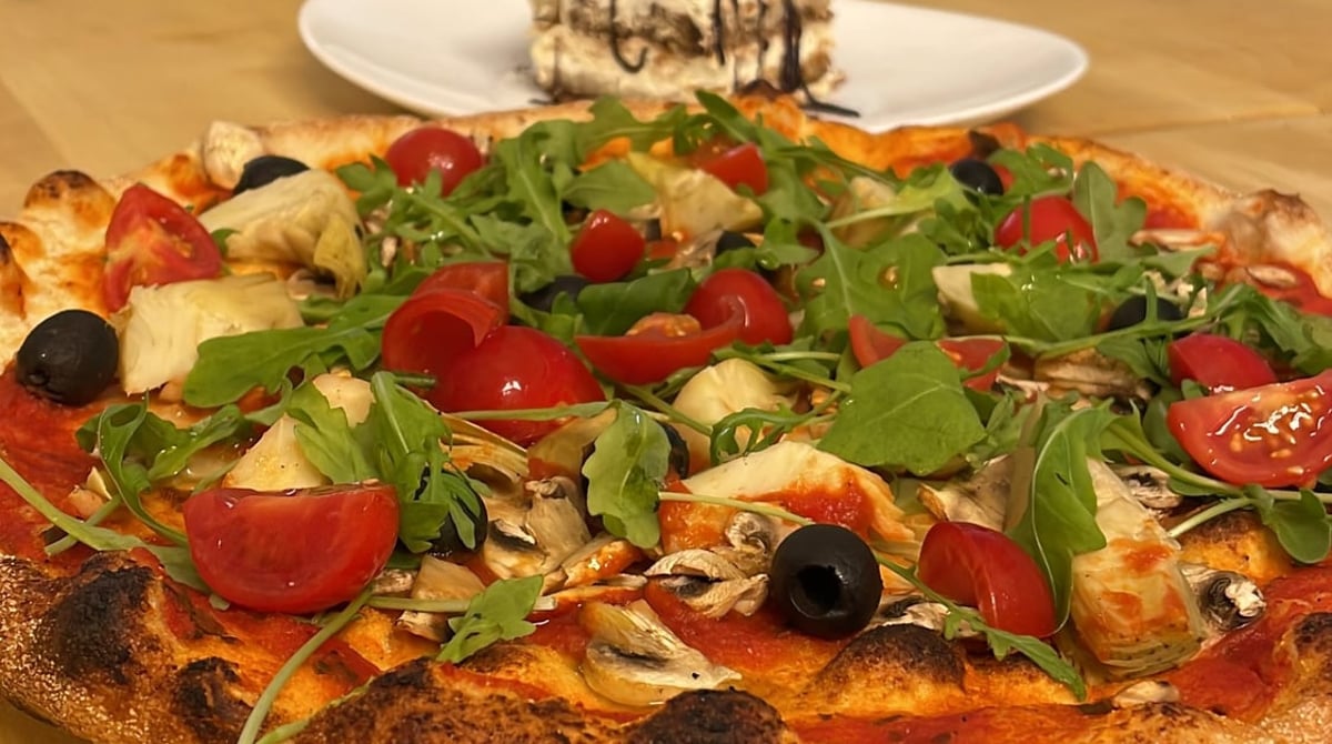 Pizza mit Rucola, Kirschtomaten, Artischocken und Oliven, dahinter ein Tiramisu, in der Pizzeria La Migliore in München-Giesing