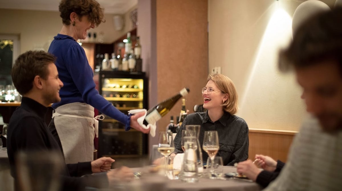 Gastraum des Zauberberg mit Servicekraft, die einem Gast Weißwein einschenkt, im Hintergrund Weinregal und Bar