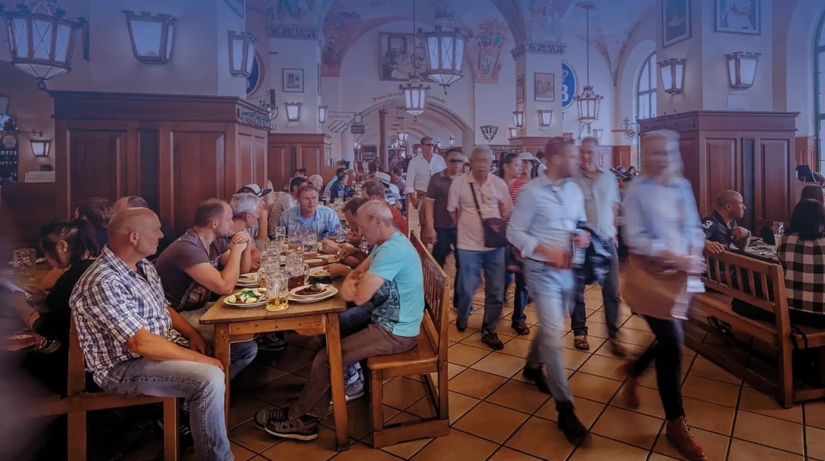 Gäste an den langen Tischen der Schwemme im Hofbräuhaus, davor Besucher im Gang