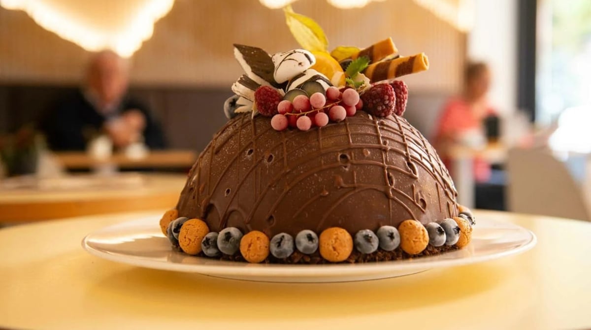 Schokolierte Eisbombe mit Beeren, Waffelröllchen und Keksen auf einem Teller, im Hintergrund der Gastraum des Eiscafés