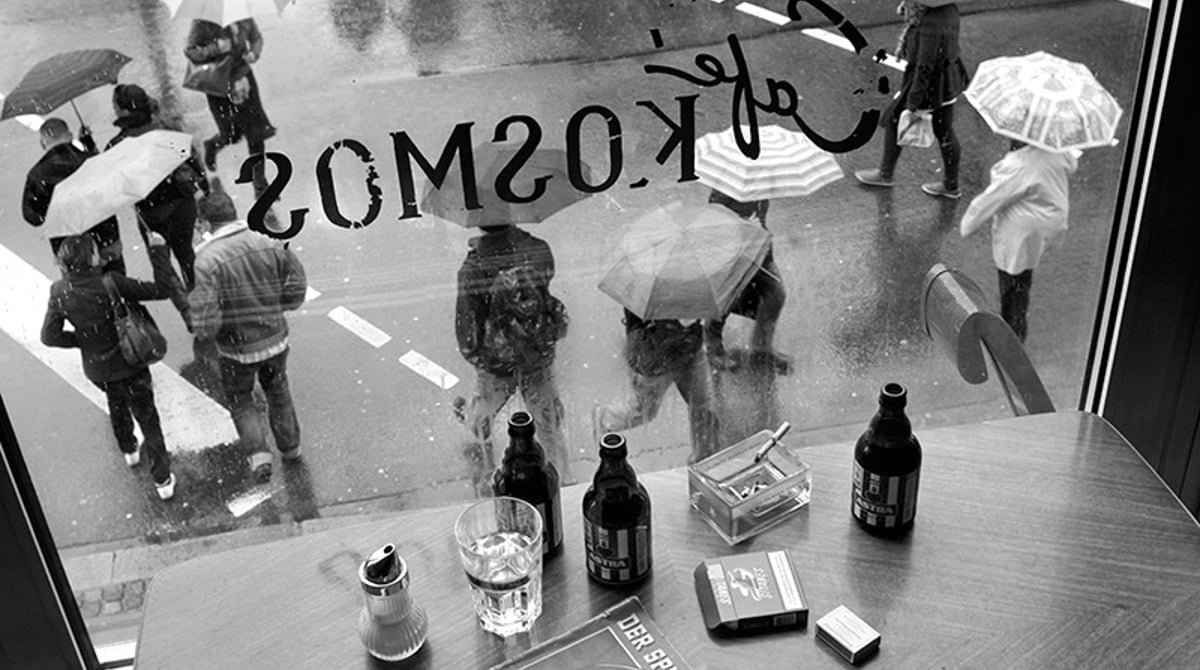 Blick durch das Schaufenster des Café Kosmos mit gespiegeltem Schriftzug auf Passanten mit Regenschirmen, davor ein Tisch mit Flaschen und Aschenbecher