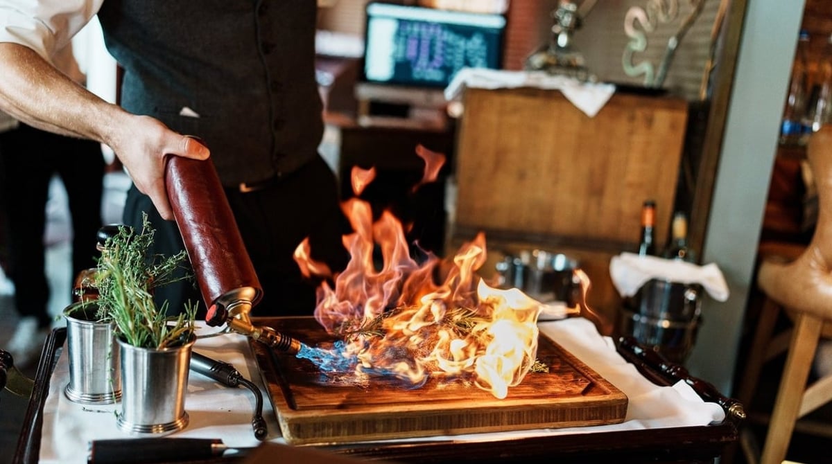 Kellner flambiert ein Steak auf einem Holzbrett mit Rosmarin am Tisch