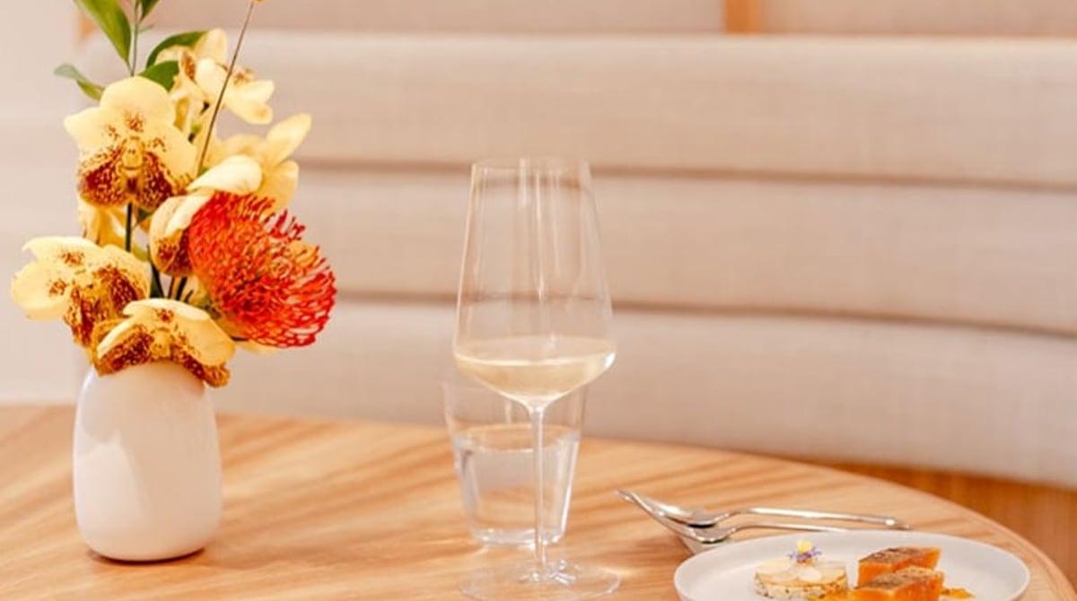 Gedeckter Tisch im KOMU mit Blumenvase, Weißweinglas und kleinem Gericht vor einer hellen Polsterbank