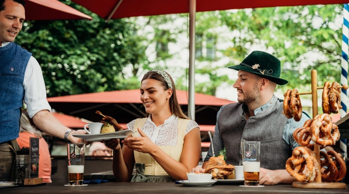 Gäste im Biergarten des Pschorr, die Bedienung bringt Speisen, daneben ein Brezenbaum