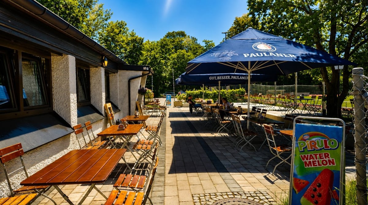 Biergartenterrasse mit Holztischen und blauen Sonnenschirmen vor dem Gebäude des Perlacher Bachgartens