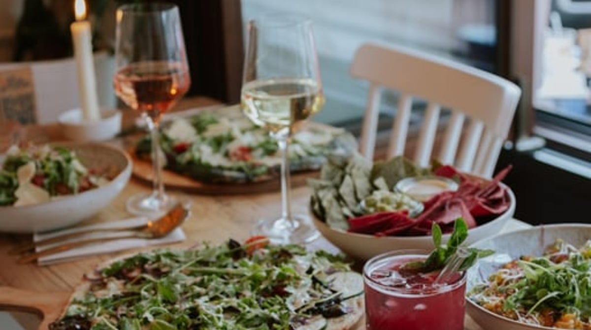 Holztisch am Fenster mit Flatbreads, Salaten, Rosé und rotem Getränk