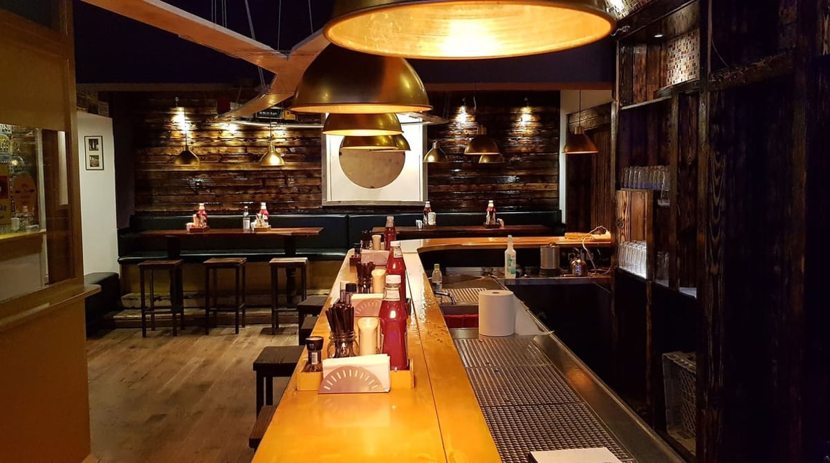 Gastraum von Taste my Burger mit langem Holztresen, Barhockern, Holzregalen und warmen Hängeleuchten