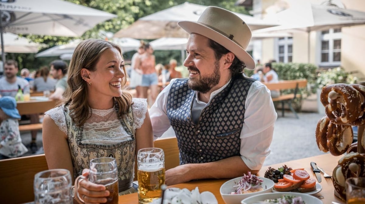 Eine Frau und ein Mann in Tracht sitzen im Biergarten an einem gedeckten Tisch und lächeln