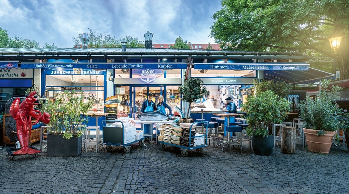 Stand von Fisch Witte auf dem Viktualienmarkt mit blauer Fassade, Bistrotischen, Pflanzen und einer roten Hummerfigur am Abend