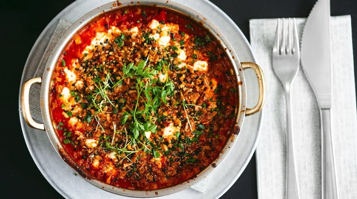 Shakshuka mit Feta und Kräutern in der Pfanne, daneben Besteck