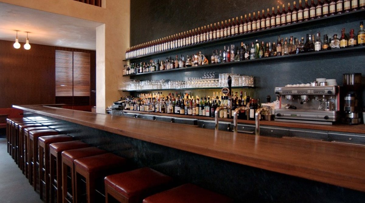 Lange Holzbar mit roten Barhockern und einem Regal voller Flaschen in Schumann's Bar