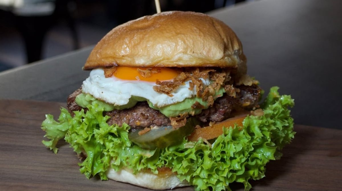 Burger mit Spiegelei, Röstzwiebeln, Avocadocreme und Salat auf einem Holzbrett