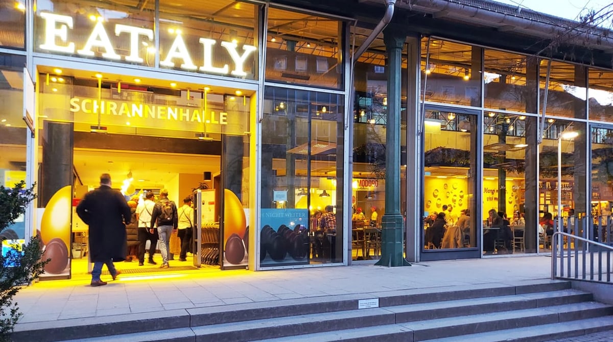 Eingang von Eataly in der Schrannenhalle am Viktualienmarkt mit leuchtendem Schriftzug und Glasfassade in der Abenddämmerung