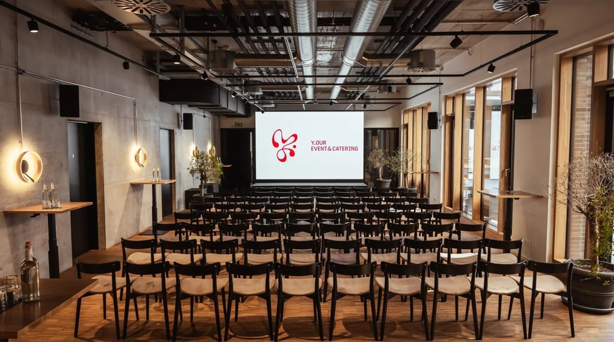 Heller Saal des Y.OUR Pavillons mit Reihenbestuhlung, Betonwänden, Parkett und hohen Fenstern