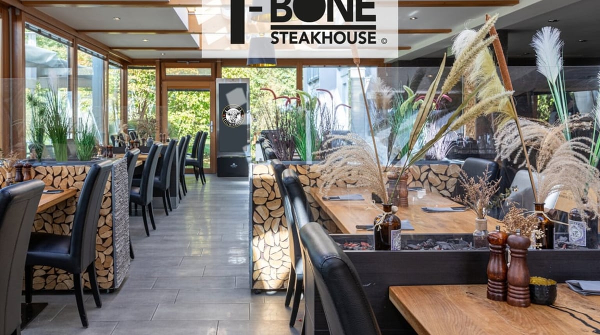 Gastraum des T-Bone Steakhouse mit Fensterfront, dunklen Tischen und Holzwand