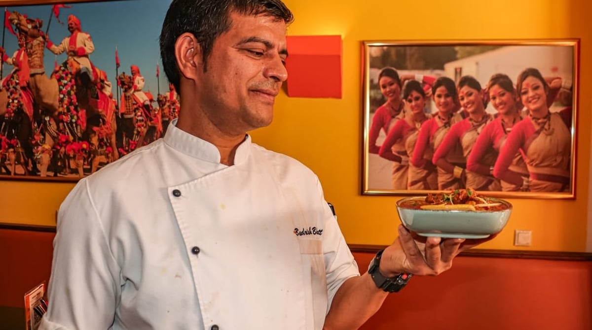 Koch mit einer Schale in der Hand vor einem Wandbild mit Tänzerinnen im Indian Mango