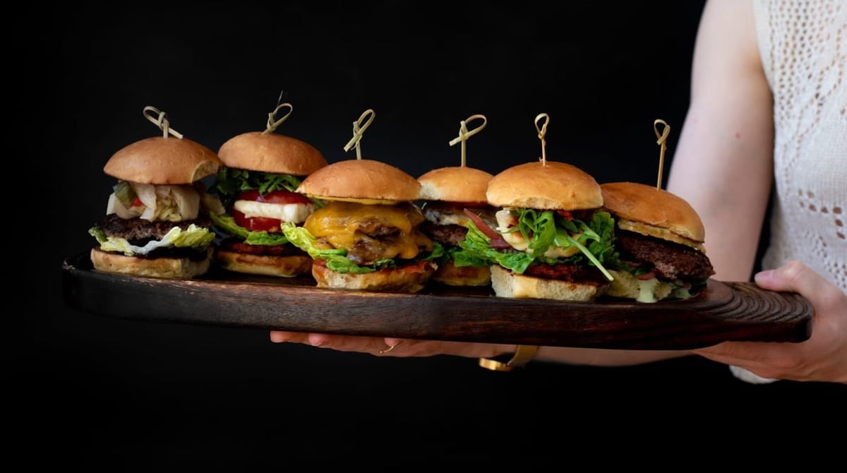 Holzbrett mit sechs Mini-Burgern, gehalten vor dunklem Hintergrund