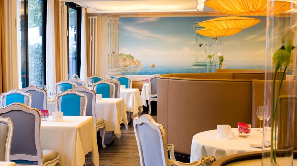 Gastraum des Restaurants Acquarello mit gedeckten Tischen, türkisfarbenen Stühlen und gemalter Küstenlandschaft an der Wand