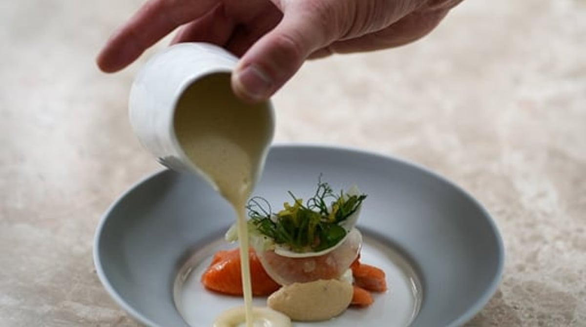 Eine Hand gießt am Tisch eine helle Sauce in einen Teller mit Gemüse und Kräutern