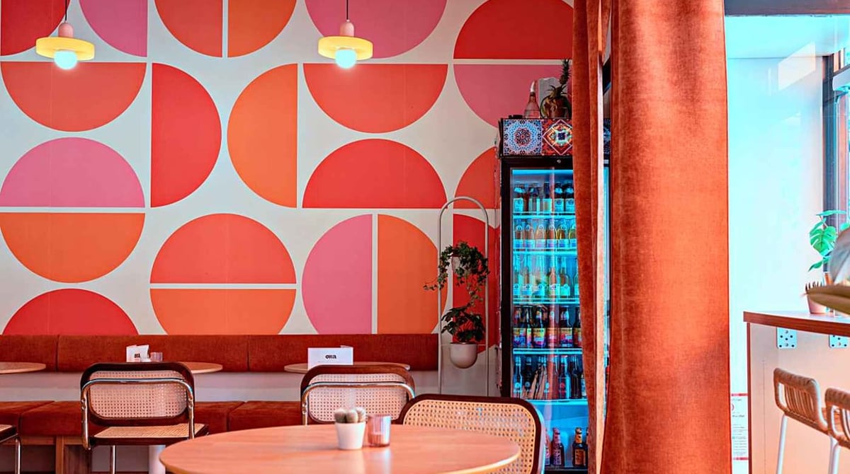 Gastraum der ORA Tagesbar mit Retro-Tapete in Pink und Orange, orangem Vorhang, Rattanstühlen und rundem Holztisch