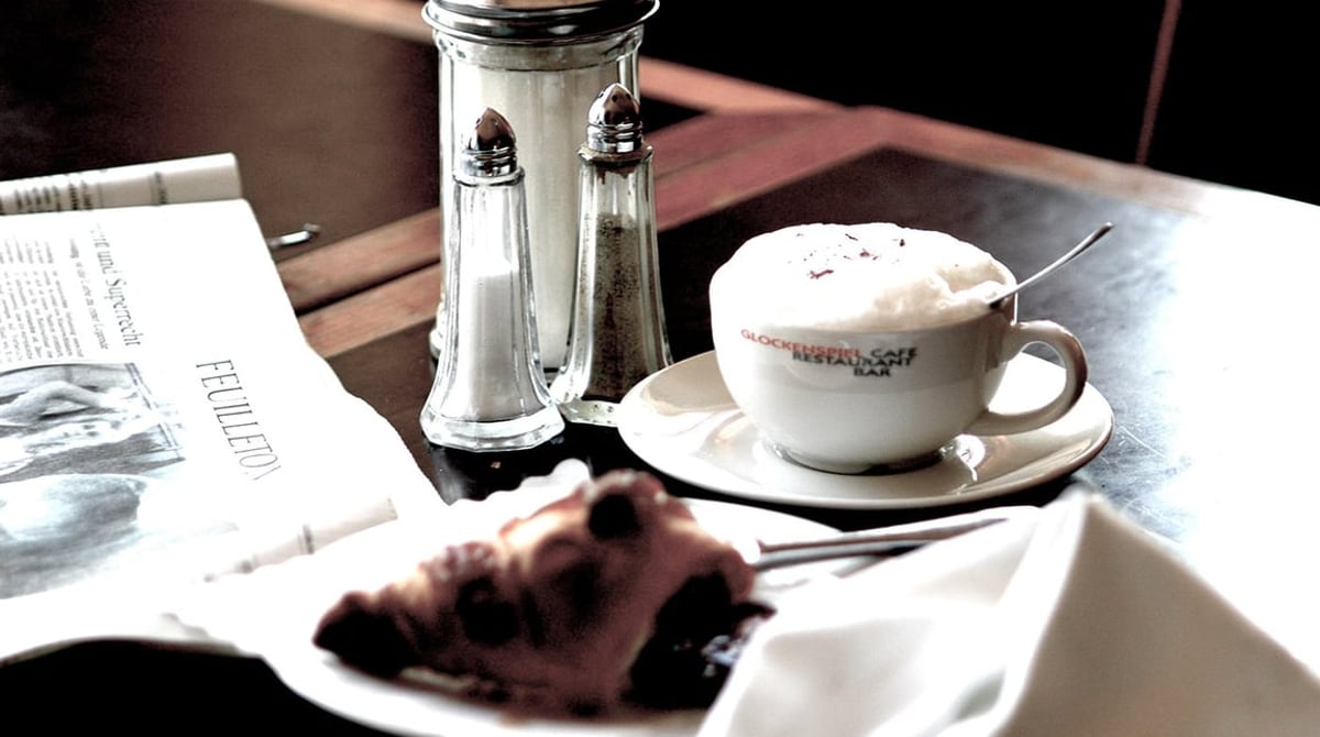 Cappuccino und ein Stück Kuchen auf einem Tisch, daneben Zuckerstreuer und Zeitung