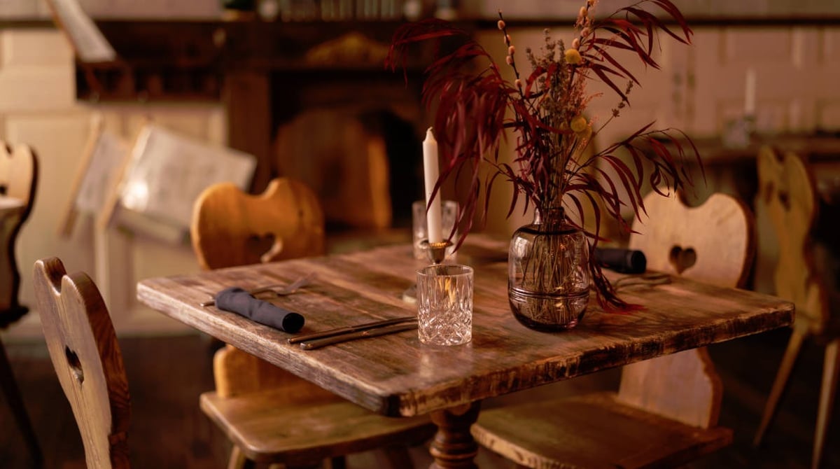 Gedeckter Holztisch mit Kerze und getrockneten Gräsern in der Vase, im Hintergrund Holzstühle mit Herz-Ausschnitt