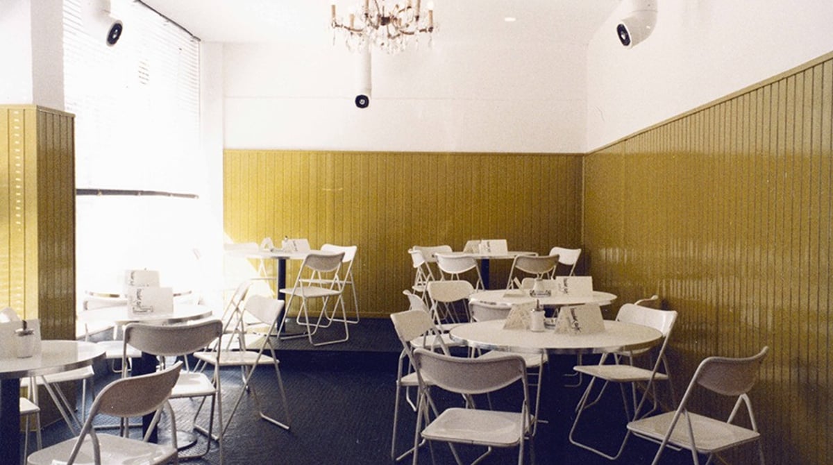 Der Gastraum des Baader Cafés mit gelb gestrichener Holzvertäfelung, weißen Klappstühlen, runden Tischen und einem Kronleuchter