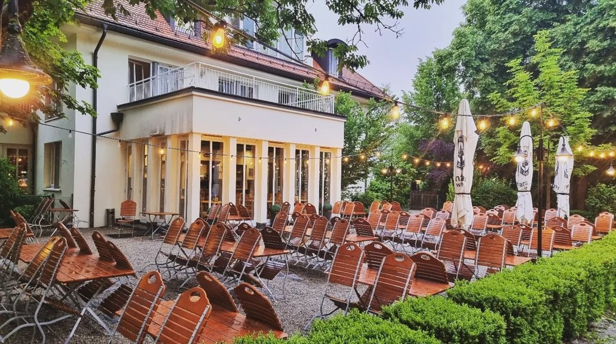 Haus mit Biergarten in der Dämmerung, davor Holztische und Stühle unter Lichterketten und Bäumen, Gasthaus Maxhof in München