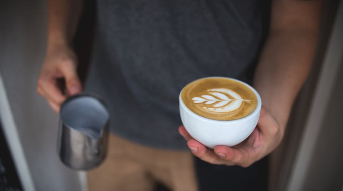 Hände halten eine Tasse Cappuccino mit Latte-Art-Blatt und eine Milchkanne aus Edelstahl