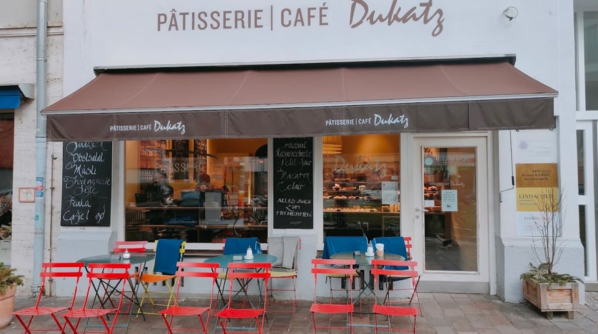 Ladenfront der Pâtisserie Café Dukatz mit brauner Markise, Kreidetafeln und roten Klappstühlen vor dem Laden