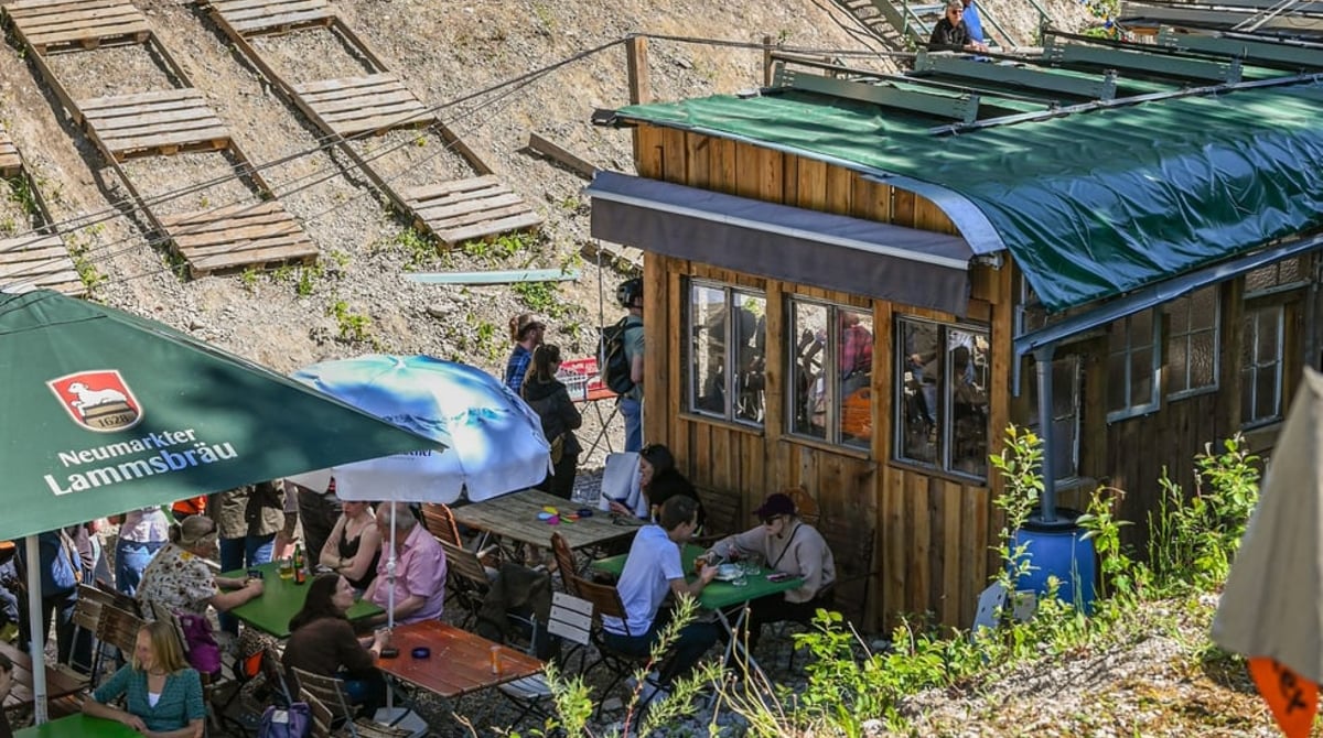 Gäste an Biergartentischen unter Sonnenschirmen vor der Holzhütte, dahinter der Kieshang der Baugrube
