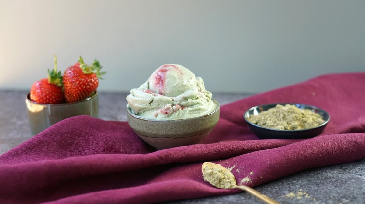 Pistazieneis mit Erdbeerswirl in einer Schale, daneben frische Erdbeeren und Pistazienpulver auf einem roten Tuch