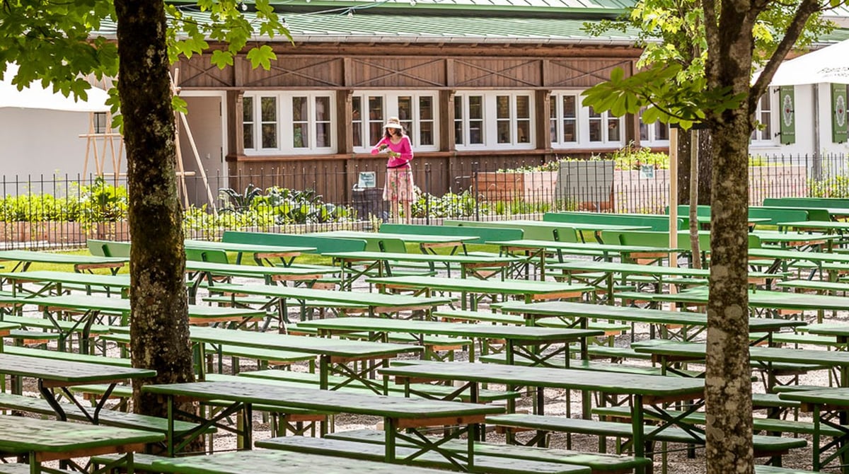 Grüne Bierbänke unter Bäumen im Biergarten der Kugler Alm, dahinter das Holzhaus mit Kräutergarten