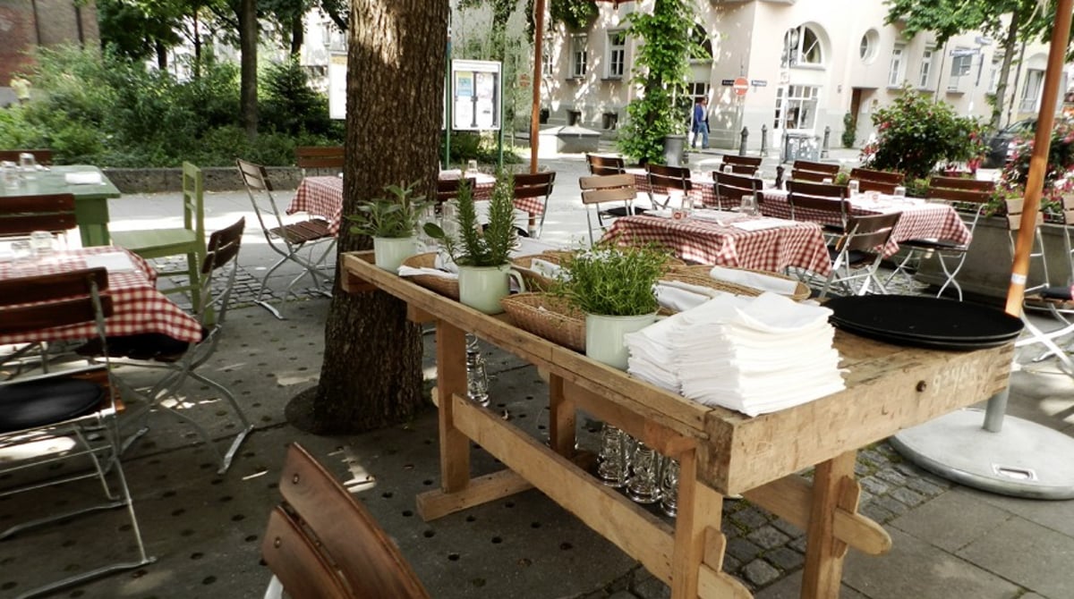 Terrasse des Chez Fritz unter Bäumen mit rot-weiß karierten Tischdecken und Sonnenschirmen