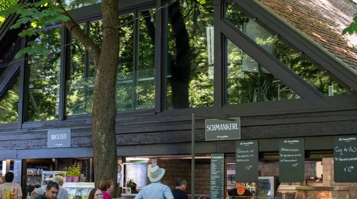 Hütte mit Dachgiebel unter hohen Bäumen, davor besetzte Biergartentische