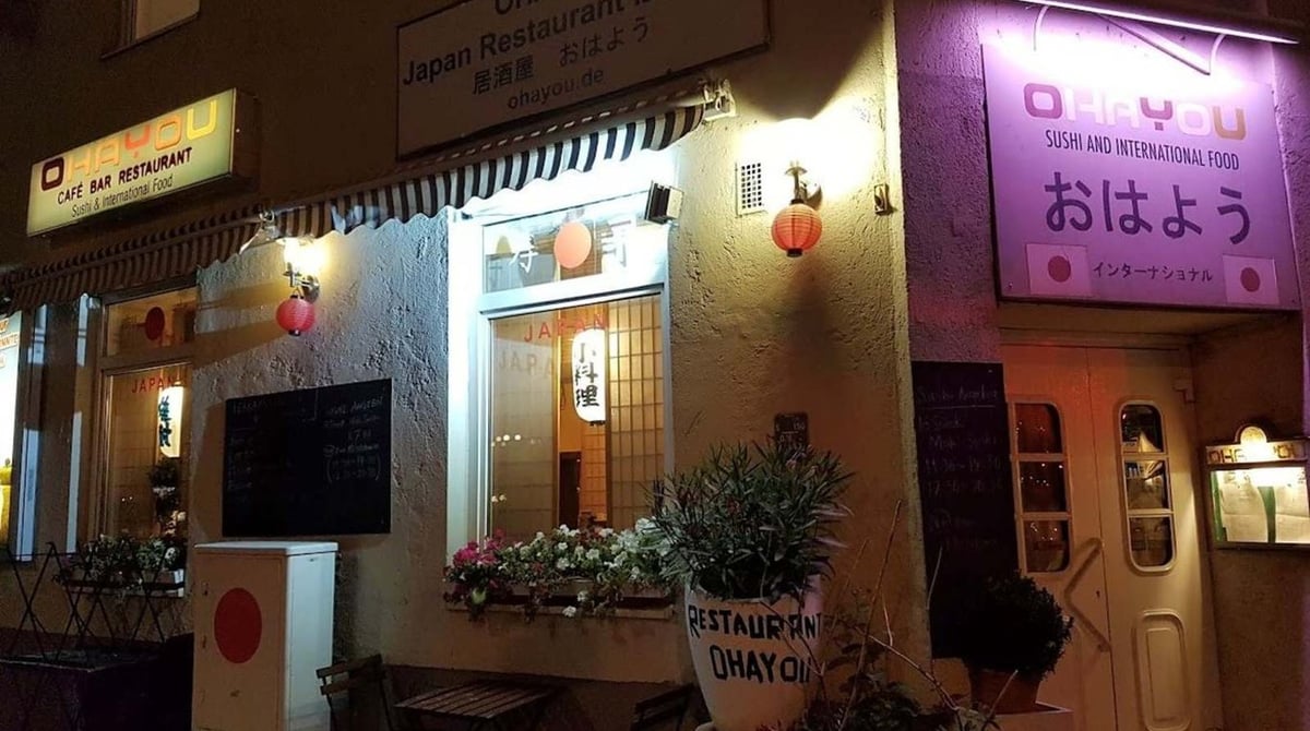 Außenansicht des Ohayou am Abend mit beleuchteten Schildern, Papierlaternen, Blumenkasten und Eingangstür
